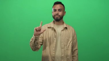 İşaretli gömlek giyen adam, stüdyoda parlak yeşil ekran ve nötr ifade ile işaret parmağı kameraya; şüphecilik.
