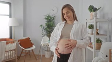 Bebek odasında hamile bir kadın karnını nazikçe tutuyor. Bebek odası dekoruyla çevrili. Huzurlu bir evde annelik beklentilerini dile getiriyor..