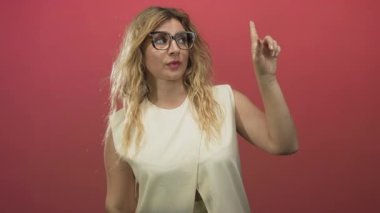 Gözlük takan sarışın bir kadın stüdyoda parmaklarını yukarı kaldırarak pembe duvarlı, dalgalı saçlı, kolsuz bluzlu, kendine güvenini güçlendiren bir kadın..