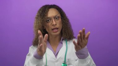 Kıvırcık saçlı, kaşları çatık kadın doktor avuçlarını öne uzatıyor ve mor stüdyoda steteskop takıyor..