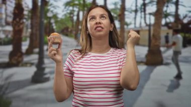 Kadın kek tutuyor, yumruğunu sıkıyor ve yakındaki sokak lambaları ve yayalarla dolu bir palmiye ağacına bakıyor..