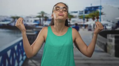 Deniz kenarındaki gezinti alanında yeşil askılı tişört ve gözlük takan kadın parmaklarını çimdikliyor; dinginlik farkındalığı.