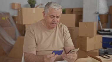 Elinde kredi kartı ve akıllı telefon bulunan adam, bir binada taşınan kutular arasında masada otururken kartviziti telefonla karşılaştırıyor; maddi kaygılar.