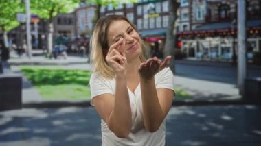 Kadın, genç sarışın, çimdikleyen parmaklar ve güneşli bir şehir sokağında kameraya doğru avuç içi açık tutmak; neşeli bir davet.