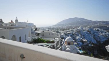 Santorini yamaç kasabası, yumuşak bokeh, uzak beyaz binalar ve ötesinde bulanık deniz, güneşli teras ve ön plandaki Wall Street ile dağılmış..