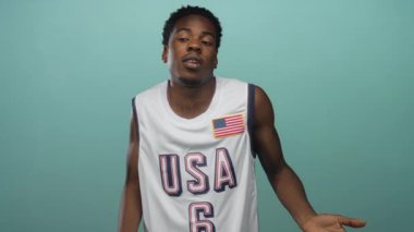 ABD basketbol formalı genç Afro-Amerikan bir adam açık elle deniz mavisi duvarına karşı omuz silkiyor..