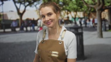 Yeşil ağaçlarla çevrili, kahverengi önlüklü kadın gündüz vakti arka planda bulanık yayalar görüyor..