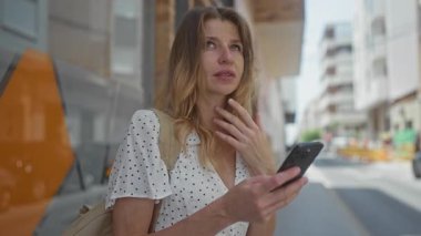 Şehir caddesinde, dükkan penceresinin yanında elinde akıllı telefon tutan kadın yukarı bakıyor ve bekliyor..