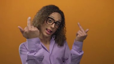 Gözlük takan bir kadın stüdyoda orta parmak hareketini turuncu bir arka plana karşı elleri ve dili açık gösteriyor..