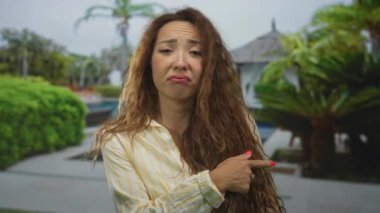 Kadın otelin havuz kulübesini işaret ediyor, yüzünde sırıtış ve el açıkça görülüyor bir otelde dışarıda dururken; oyuncu bir güven..