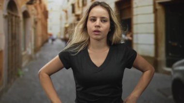 Dar bir sokakta elleri kalçasında, kapalı gözleri olan, siyah v yaka tişört giyen ve sıradan duruş takınan genç sarışın bir kadın..