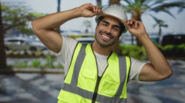 Adam, palmiye ağaçları ve park halindeki arabalarla kaplı kentsel caddede yansıtıcı yelek giyerken beyaz kaskı ayarlar; güvenlik garantisi.