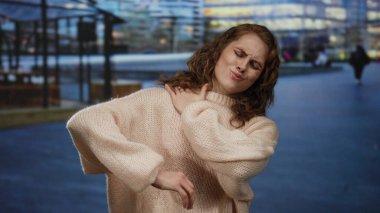 Gün batımında şehir merkezinde süveter giyen bir kadın, bulanık ışıklarla dolu bir şehir ortamında omzuna dokunurken rahatsızlığını dile getiriyor..