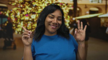 Mavi gömlek giyen Afro-Amerikalı kadın el sallıyor ve sokak pazarında parmağını gösteriyor; dostça karşılama neşe sıcaklığı.