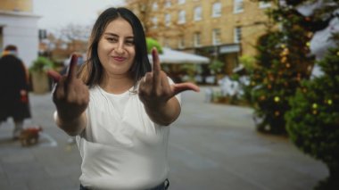 Genç İspanyol kadın şehir caddesinde dekore edilmiş saksı ağaçları ve tuğla bina cepheleriyle orta parmağını kameraya doğru gösteriyor..