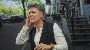 Kısa saçlı bir kadın, gün ışığında irkilirken kulağına dokunur. Hayal kırıklığı, acı, işitme duyusu..