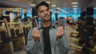 Genç İspanyol bir adam spor salonunda, spor aletleri ve tavan lambalarıyla çevrili bir şekilde para jesti yapıyor. Bu da spor ortamında motivasyon ya da finansal hırs olduğunu gösteriyor..