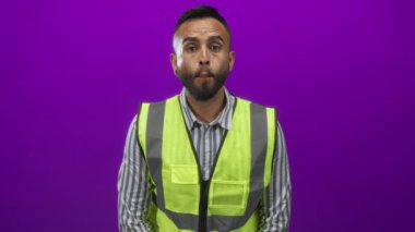 Yansıtıcı güvenlik yeleği ve çizgili gömlek giyen bir adam stüdyo mor arka planına bakarken dudaklarını büzüştürüyor..