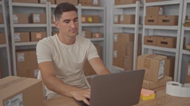 Adam dizüstü bilgisayarda elleri klavyede karton kutular ve paketlerin arasında yazıyor; küçük işletme kararlılığı.