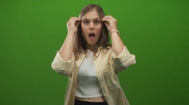 Genç sarışın kadın, parlak yeşil stüdyo setinde kameraya bakarken elleri ile yüzünü çerçeveler; şaşkınlık.