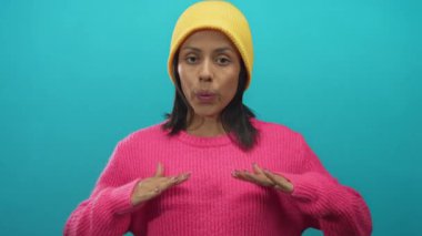 Sarı şapkalı kadın sakin bir şekilde mavi arka plana doğru meditasyon yapıyor, pembe bir kazak giyiyor ve sakin nefes alma teknikleri gösteriyor..