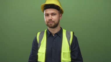 Sarı şapkalı ve yüksek görüşlü yelekli bir adam yeşil stüdyoda bana el hareketi yapıyor..