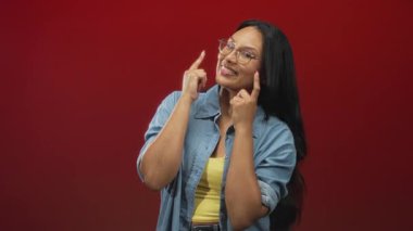 Kırmızı stüdyoda gülümseyen ve başını eğen, gözlüklü ve kot gömlekli bir kadın, neşeli bir neşe.