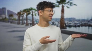 Beyaz süveter giyen ve gözlüklü adam, palmiyelerin palmiye ağaçlarının altında gezindiğini gösteriyor; belirsizlik..