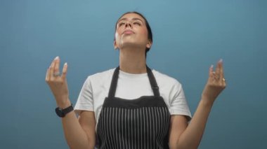 Siyah çizgili önlüklü kadın avuçlarını kaldırıp başını mavi stüdyoda kaldırıyor..