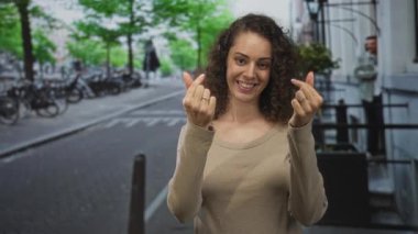 Kadın bir şehir caddesinde kameraya gülümserken iki eliyle parmak kalp hareketi yapıyor; dostça sıcak neşe.