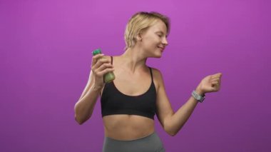 Yeşil meyve suyu şişesi tutan, çıplak karnını gösteren, elini stüdyoda kaldırmış, gülümseyen ve sallanan bir kadın; sağlık zindeliği.