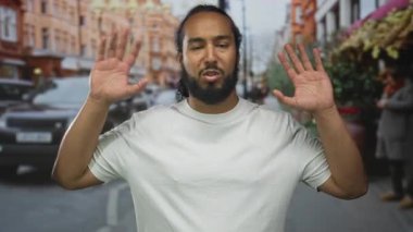 Beyaz tişörtlü ve avuç içi açık bir adam meşgul bir şehir caddesinde el hareketi, sakallı ve ilgili görünüyor; itiraz endişe.