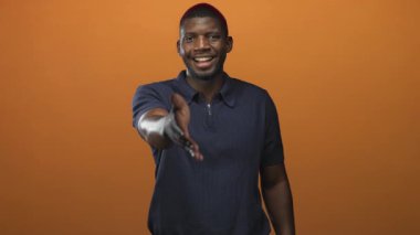 Erkek stüdyoda el sıkışmak için turuncu bir arka fona uzanır, gülümser ve hafifçe öne eğilir; dostça selamlaşır.