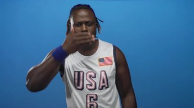 Man covers eyes with hand and raises palm in blue studio wearing usa basketball jersey and wristband; defiance resilience.