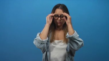 Mavi stüdyoda siyah gözlük takan ve alnına dokunan bir kadın beyaz tank ve kot gömlek giyer; yorgunluk yansıması.