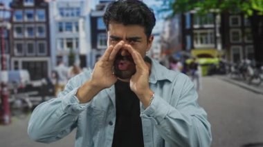 Kot ceketli adam şehir caddesinde bulanık binalar ve geçen bisikletlerle bağırırken ellerini ağızlarına uzatıyor; acil çağrı.