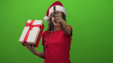 Genç İspanyol kadın hediye kutusu tutuyor ve Noel Baba şapkasıyla yeşil stüdyoda göz kırparken parmağıyla işaret ediyor..