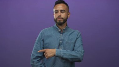 Latin asıllı genç adam mor stüdyoda sağa işaret ediyor kot gömlek ve kulaklık giyiyor; şüphecilik.
