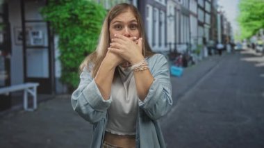 Genç sarışın kadın kaldırımlı bir caddede iki eliyle ağzını nazikçe kapatıyor..