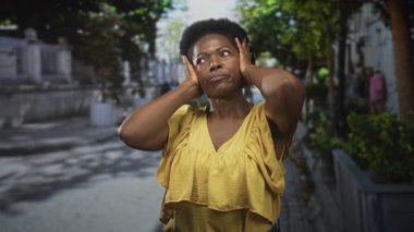 Şehir caddesinde ağaçlar ve taş ekicilerle kaplı, sarı bluz giyen, baş kenarlarını elleriyle bastıran kulakları olan bir kadın..