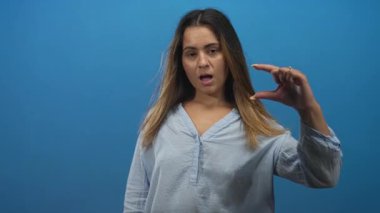 Uzun saçlı ve açık mavi bluzlu bir kadın mavi stüdyoda küçük boy göstermek için parmaklarını sağ eliyle çimdikliyor..