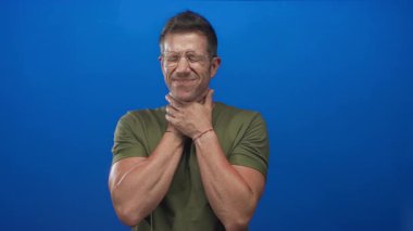 Stüdyoda boynunu tutan, gözlüklü ve zeytin gömlekli bir adam mavi arka planda boğaz ağrısı çekiyor..