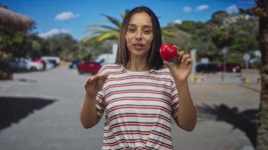 Güneşli bir sokakta palmiye ağaçları ve park halindeki arabalarla konuşurken kırmızı kalp tutan ve el kol hareketi yapan kadın. Aşk mesajı..