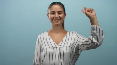 Çizgili bluzlu kadın gülümsüyor, gözleri kapalı, barış işareti ile elini kaldırıyor, soluk mavi stüdyo arkaplanına karşı, sevinç kutlaması..