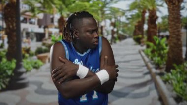 Sokakta palmiye ağaçlarıyla dolu bir sokakta kollarına basketbol forması ve bileklik takan genç adam rahatsız..