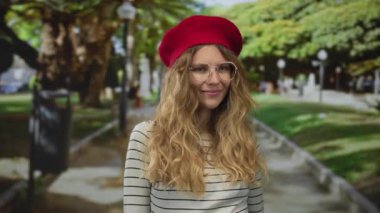 Kendine güveni tam bir kadın saçı, sarı bereli, gözlüklü ve açık havada kırmızı çizgili süveterli bir parkta gülümsüyor..