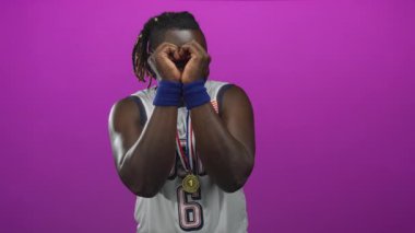 6 numaralı formayı, mavi bileklikleri ve stüdyo magenta arka planında altın madalyayı takan adam, elleri yüzün üzerinde kalp oluşturuyor..