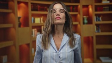 Gözlüklü ve çizgili tişörtlü genç İspanyol kadın kütüphane ahşap raflarında kollarını kavuşturuyor; reddedilme.