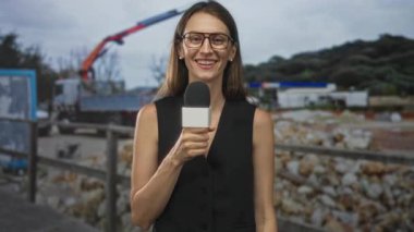 İnşaat alanında vinç ve molozların yanında mikrofon tutan kadın muhabir kameraya konuşuyor..