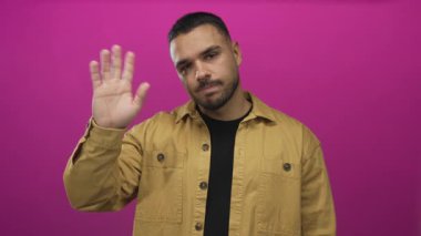 Adam stüdyoda pembe bir duvara el sallıyor, kısa kesilmiş saçlar ve ince bir gülümsemeyle; dostça bir selamlama..